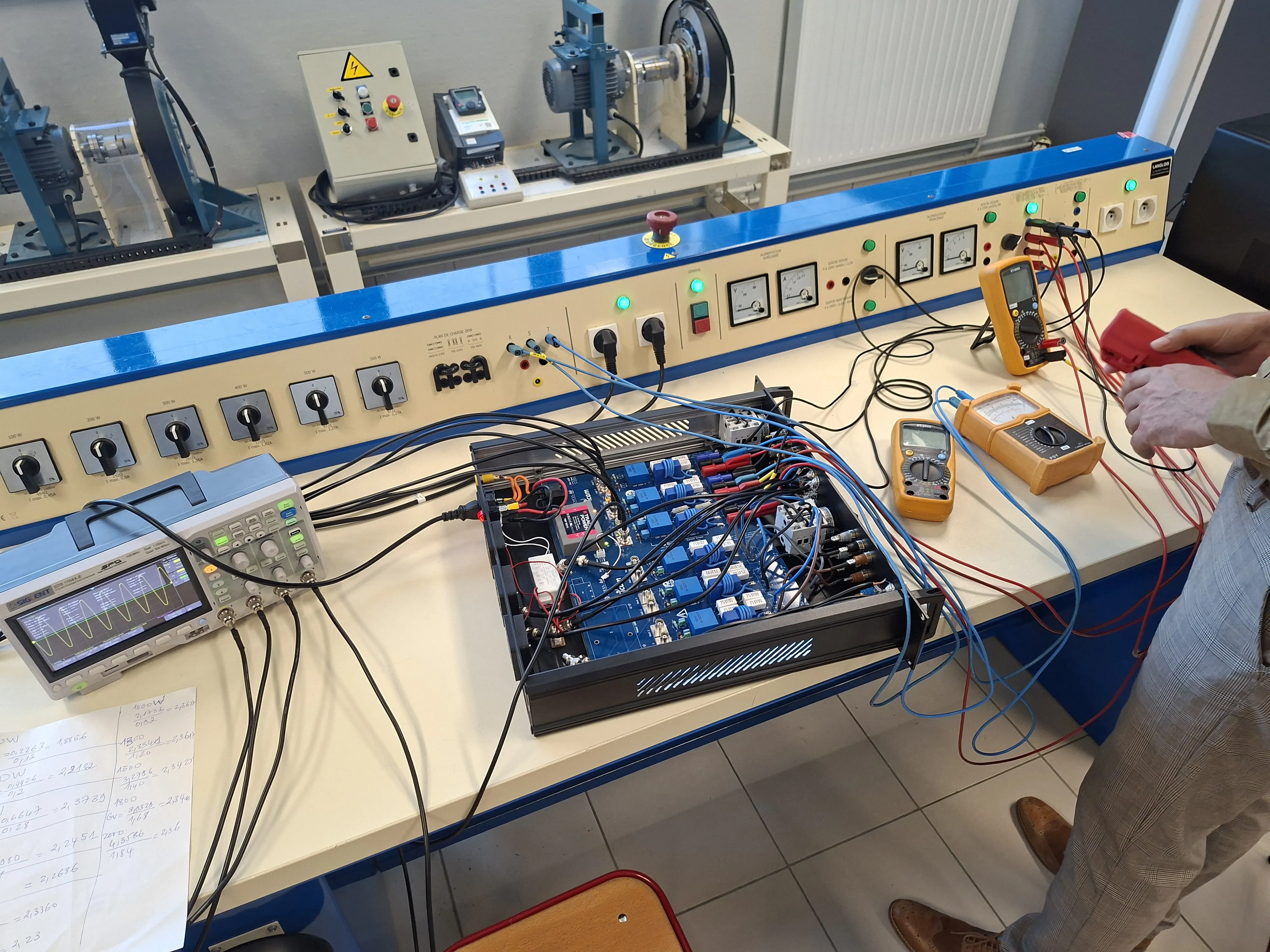 Voltage and current sensing boards in an open 19-inch chassis on a teaching bench, with gain calculations on paper